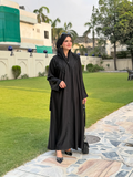 Woman in a black abaya walking outdoors on a paved path with green grass and trees in the background.