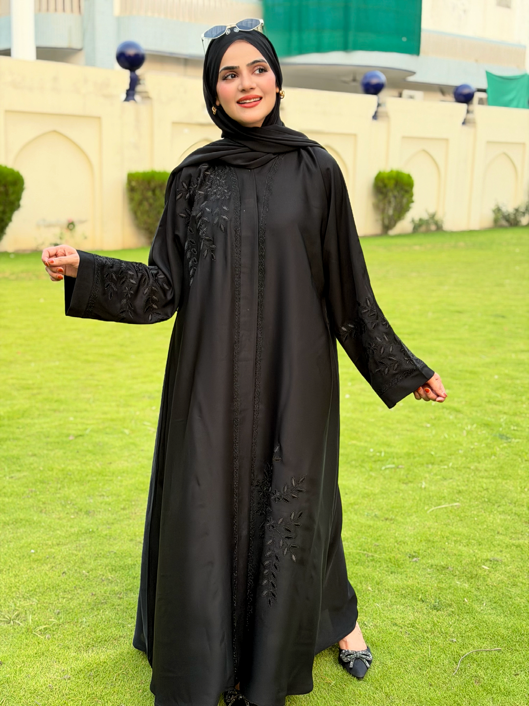 Woman wearing a black abaya with floral embroidery on a grassy area.